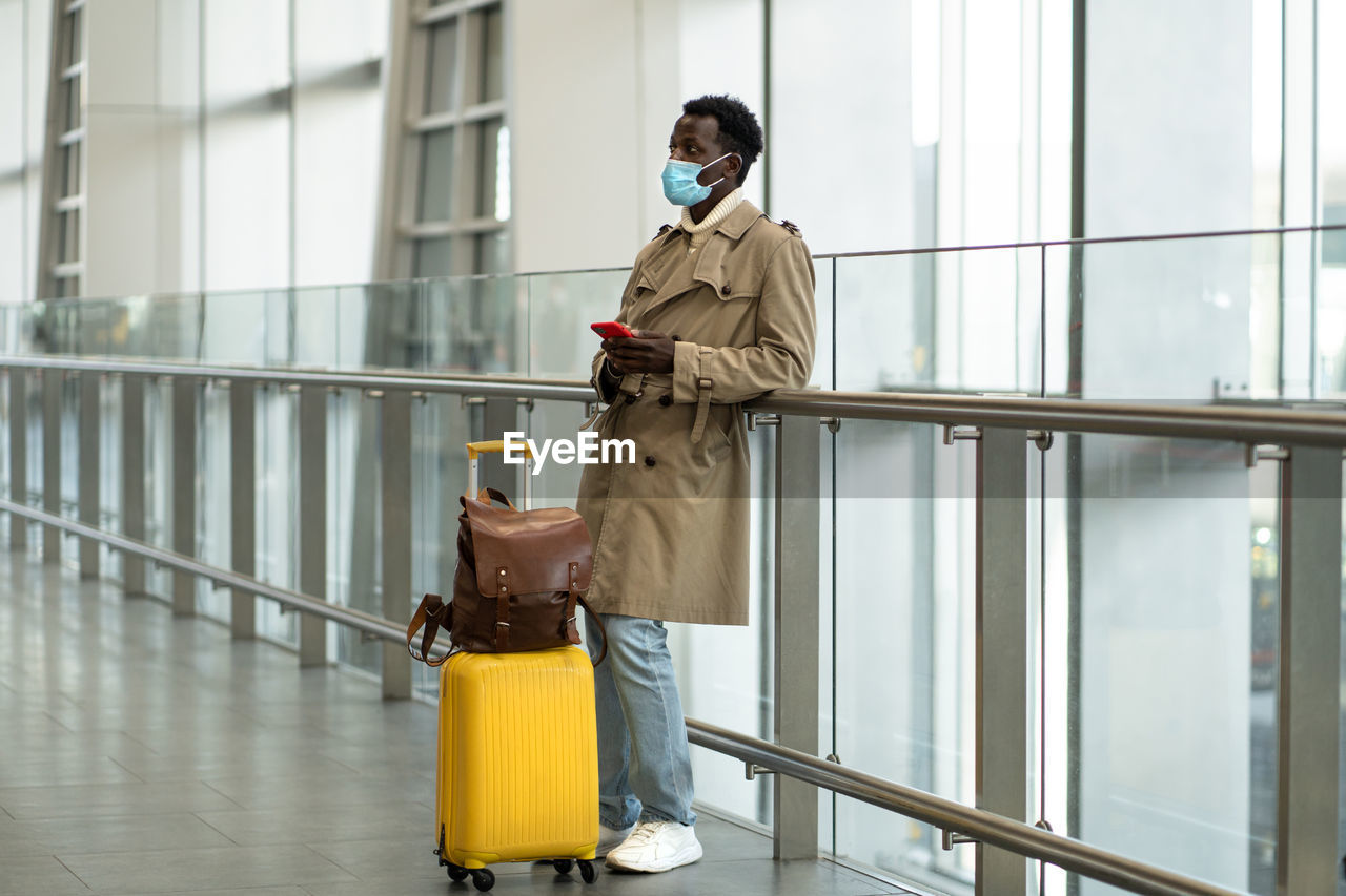 Afro traveler man stands in airport terminal, wear face mask, waiting for flight and boarding