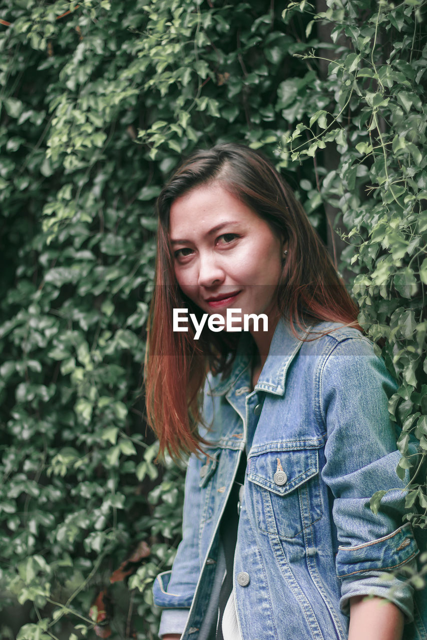 PORTRAIT OF A SMILING YOUNG WOMAN STANDING AGAINST PLANTS