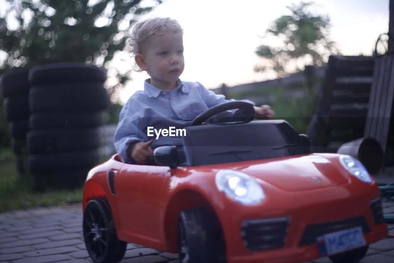 car, childhood, mode of transportation, vehicle, motor vehicle, child, transportation, one person, land vehicle, men, automobile, day, city car, portrait, front view, casual clothing, sports car, looking, sitting, toy, driving, nature, outdoors, innocence, emotion, leisure activity, standing, tree, happiness, clothing, wheel