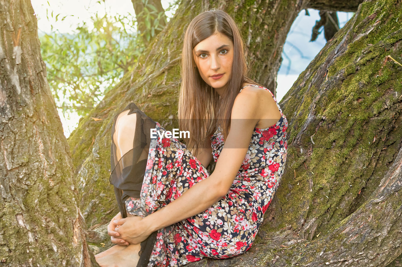 Portrait of young woman in dress sitting on tree trunk