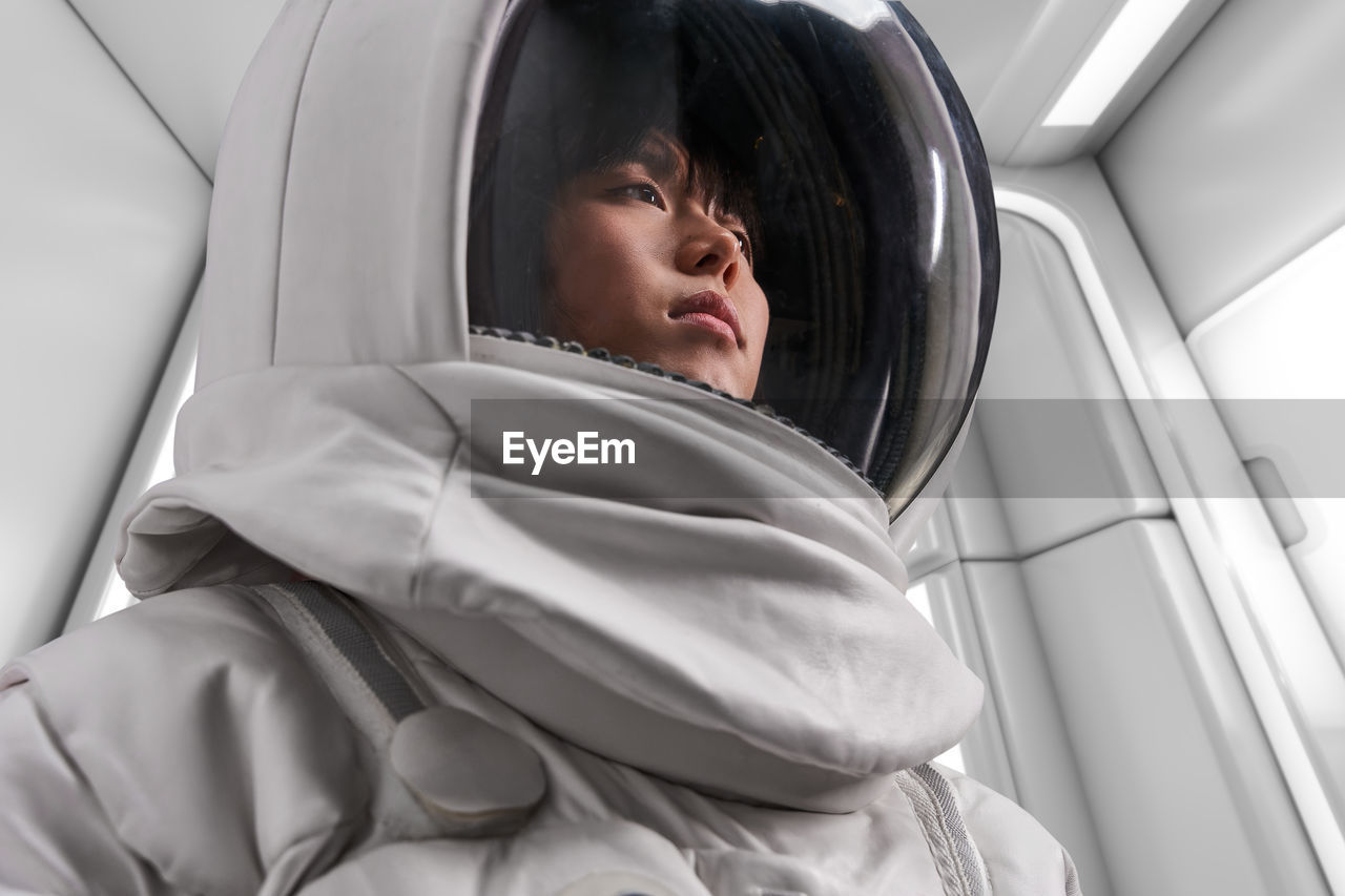 Asian woman in astronaut costume and helmet looking away during space mission in spaceship