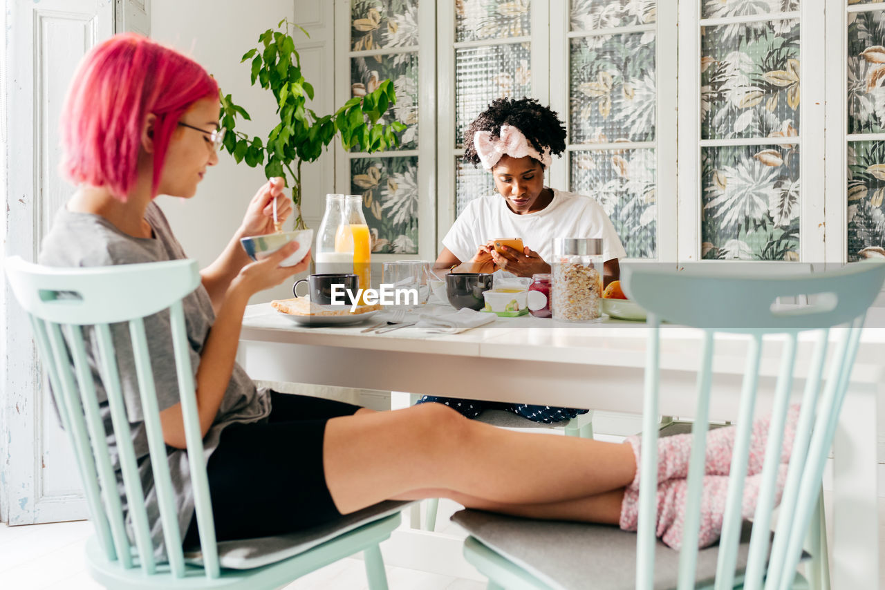 Relaxed young woman with dyed hair sitting near table and eating yummy dish near african american friend spilling cereals during breakfast in cozy apartment