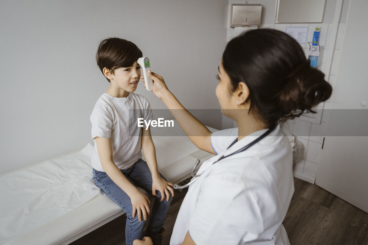 Female pediatrician measuring temperature of boy through infrared thermometer at hospital