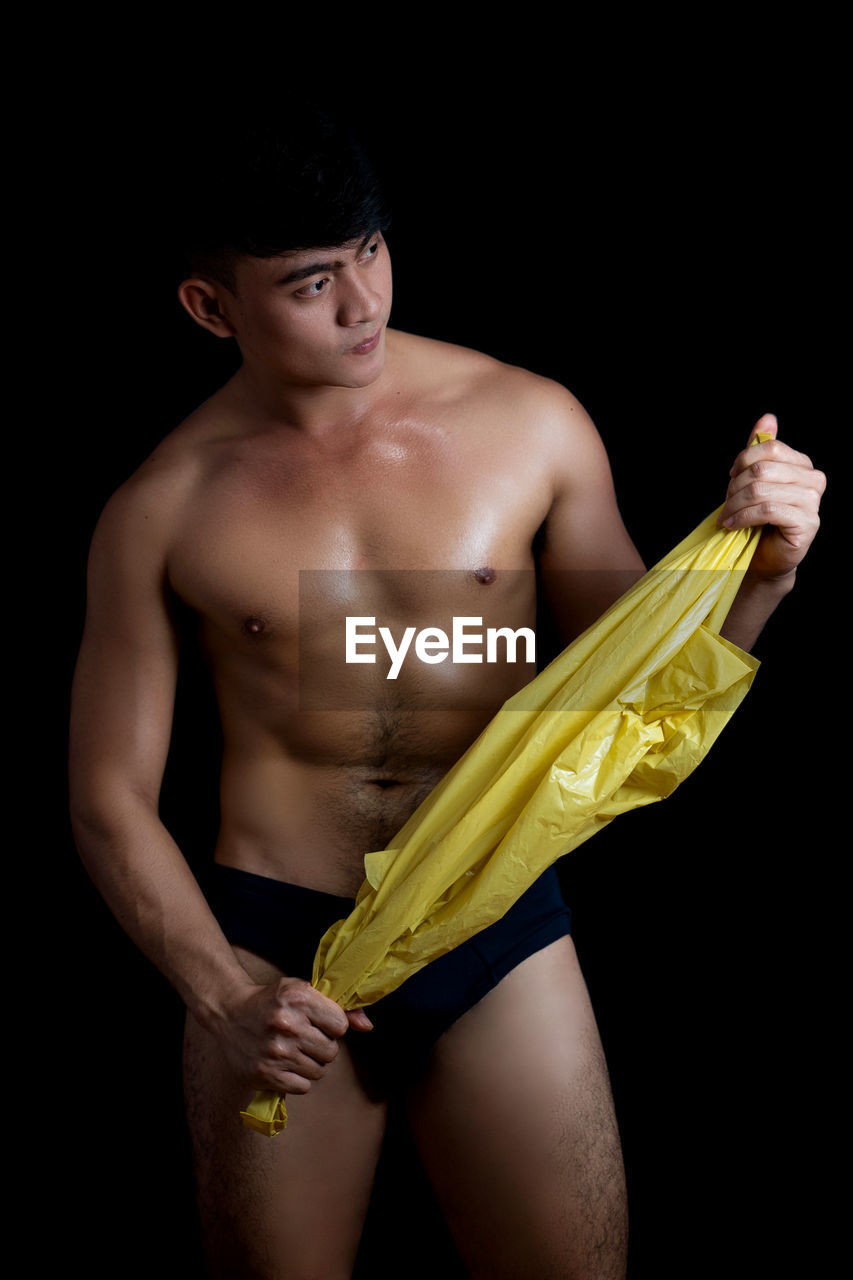 Shirtless young man holding plastic bag while standing against black background