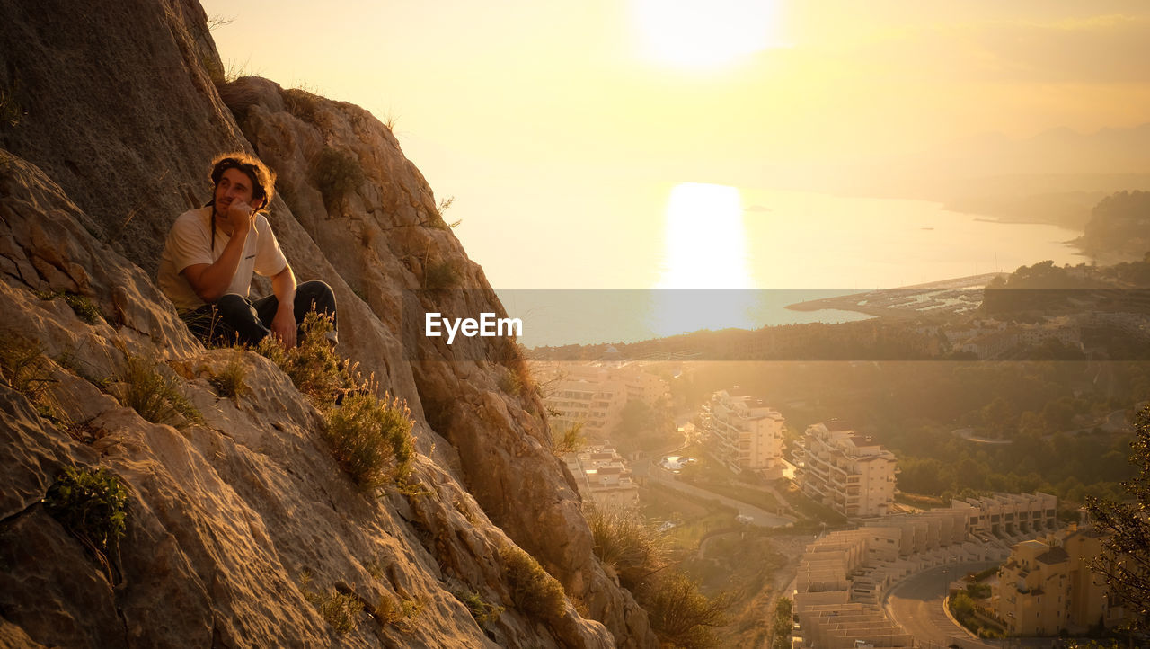 Man sitting on rocky mountain against sky during sunset