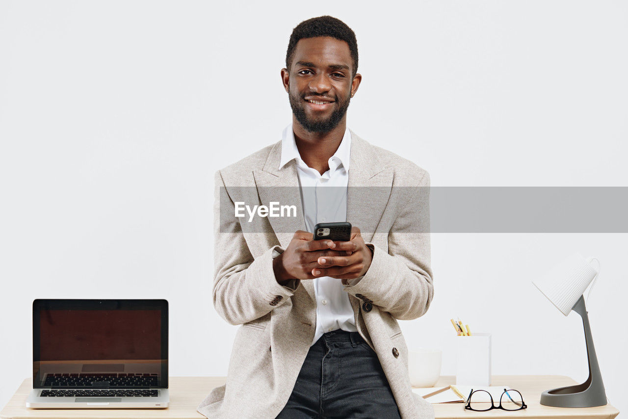 technology, wireless technology, adult, computer, one person, communication, men, business, businessman, laptop, smiling, indoors, front view, copy space, business finance and industry, conversation, happiness, using laptop, office, occupation, portable information device, portability, internet, portrait, person, table, clothing, working, computer network, beard, mobile phone, sitting, eyeglasses, smartphone, businesswear, facial hair, young adult, corporate business, white-collar worker, cheerful, glasses, looking, emotion, furniture, e-mail, holding, standing, dress shirt, looking at camera, telephone, success, place of work, desk, button down shirt, convenience, casual clothing, new business, computer equipment, professional occupation, waist up, studio shot, typing