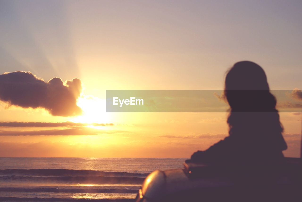 REAR VIEW OF SILHOUETTE WOMAN SITTING ON BEACH AT SUNSET