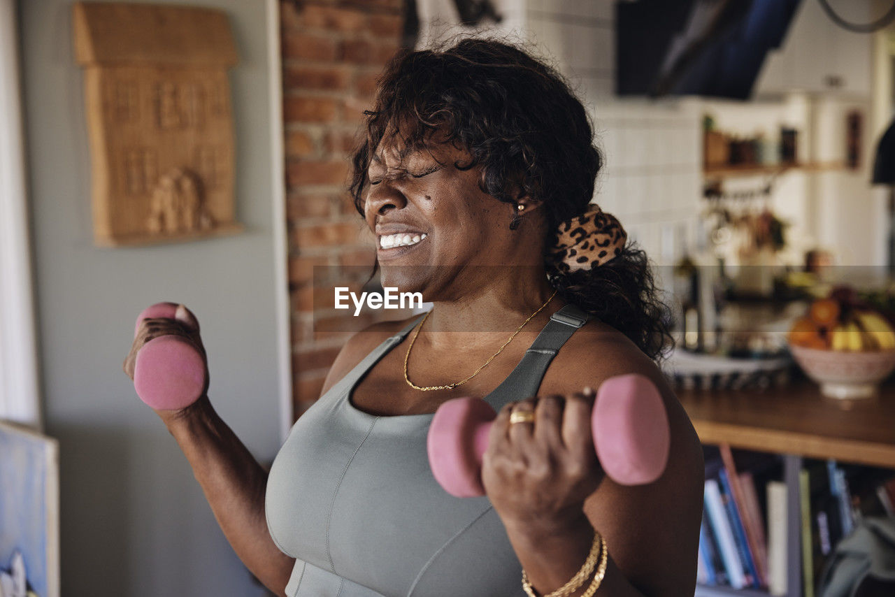Mature woman with eyes closed clenching teeth while exercising with dumbbells at home