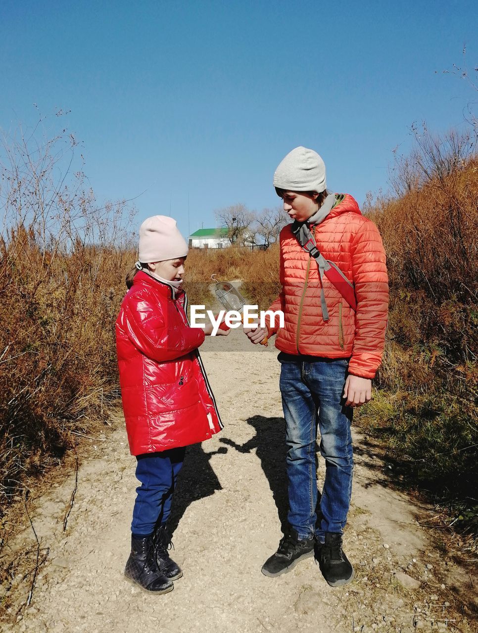 Boy and girl in red jackets holding red and orange things and standing on dirt road