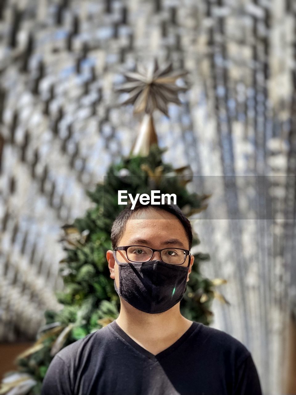 Portrait of young man wearing eyeglasses and face mask against christmas tree and skylight.