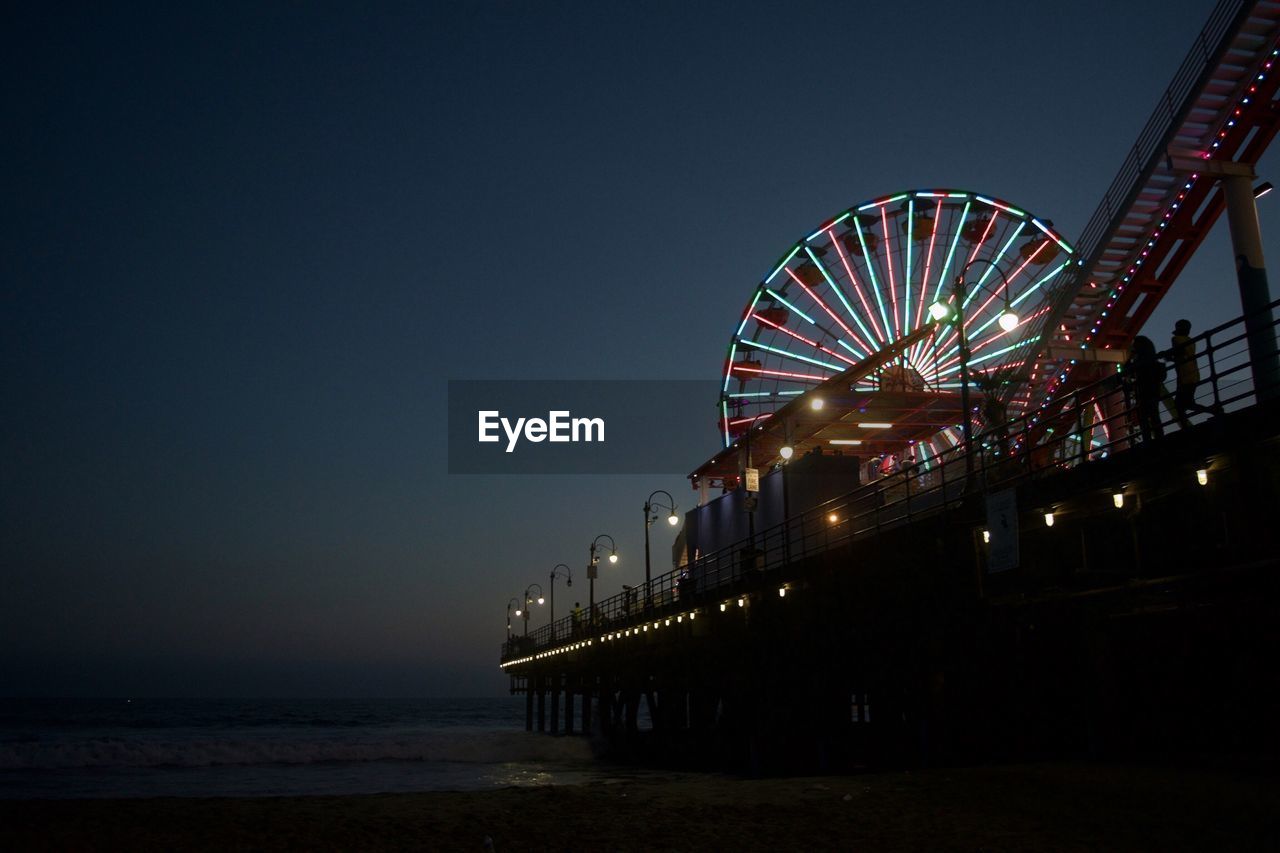 Low angle view of illuminated ferris wheel on pier by sea at night