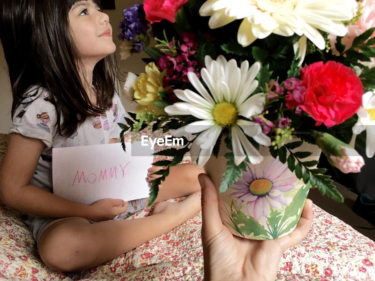 Cropped image of hand holding flower vase against girl sitting on sofa