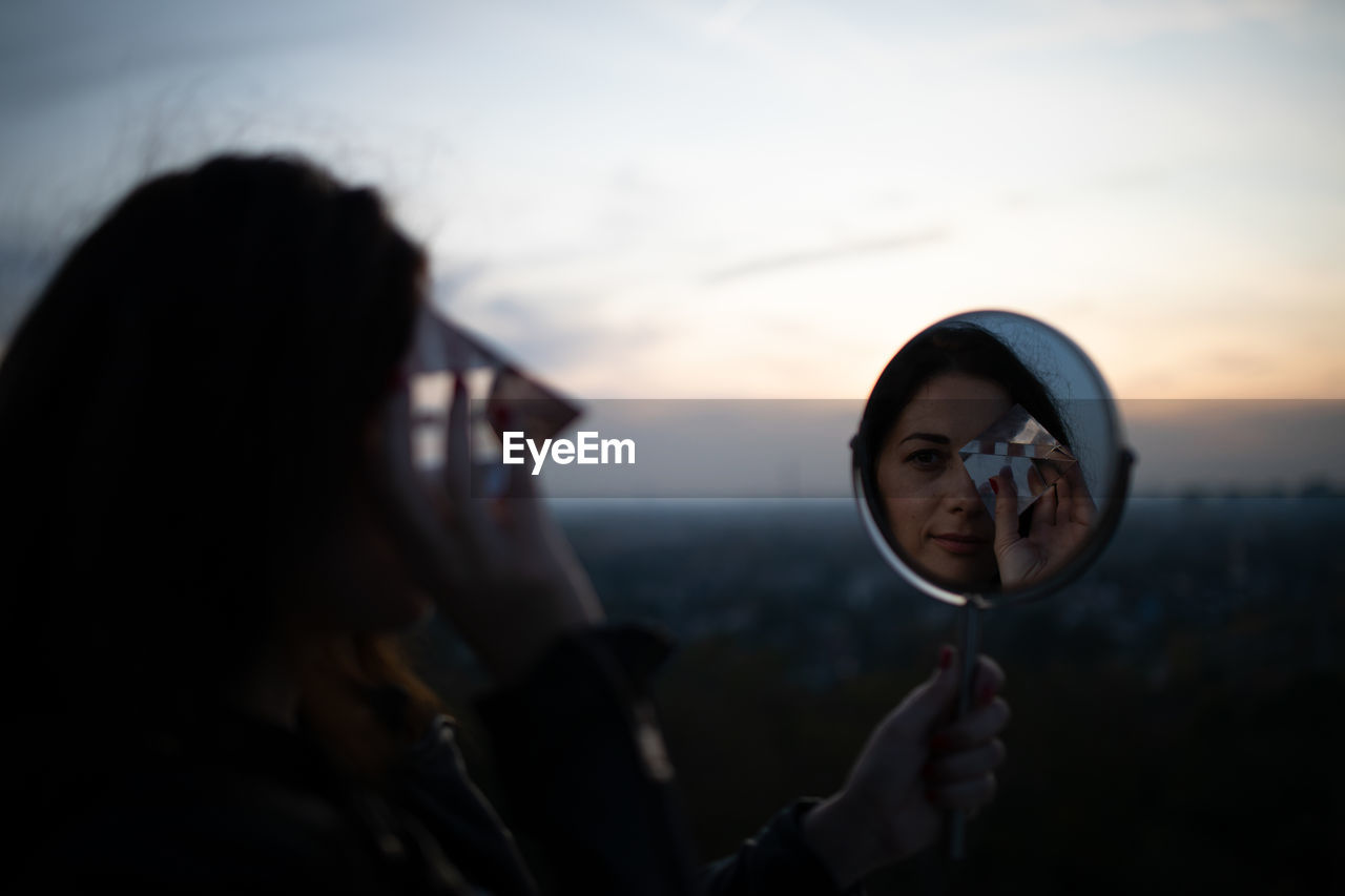 Reflection of woman holding prism in mirror against sky during sunset