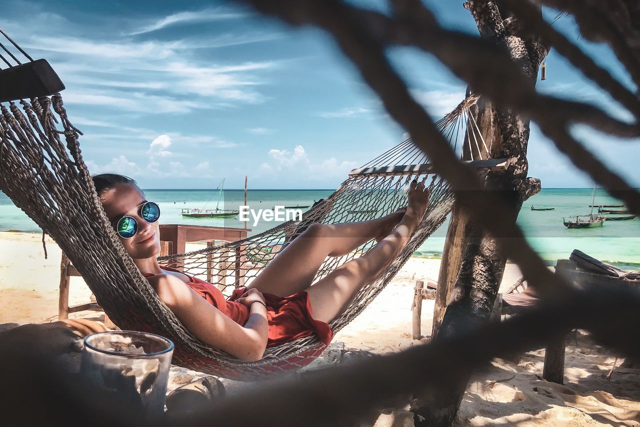 Portrait of woman relaxing on hammock at beach against sky