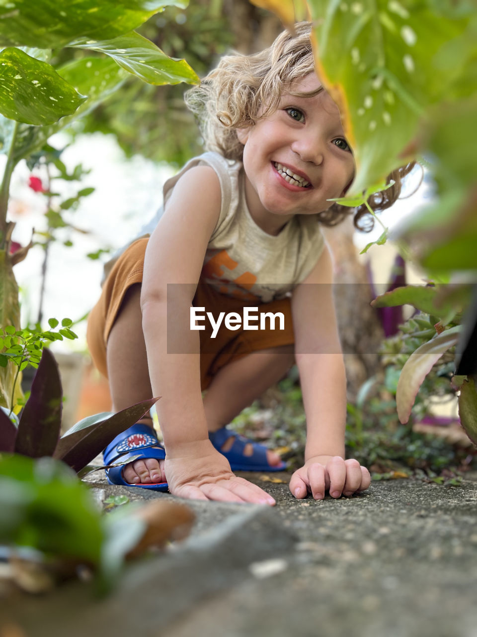 Small boy with curly hair smiling  crouched among the plants with his hands resting on the ground.