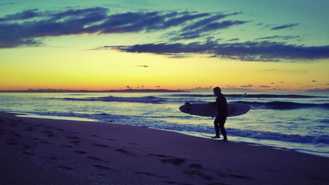 Man with surfboard walking on shore during sunset