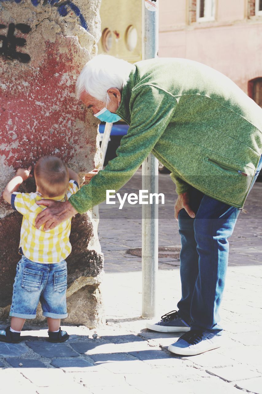 Full length of baby boy and grandfather wearing coronavirus mask  standing outdoors