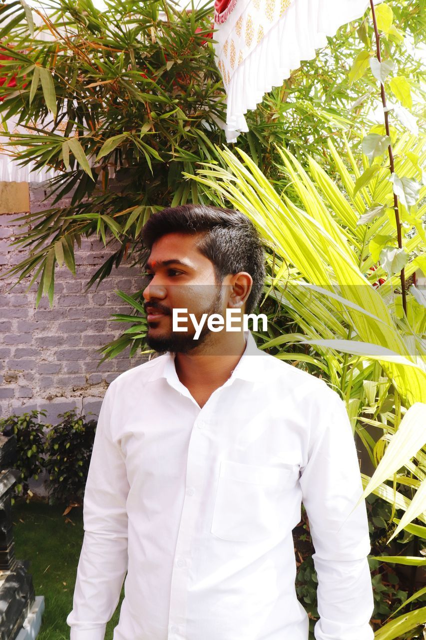 YOUNG MAN LOOKING AWAY STANDING AGAINST PLANTS