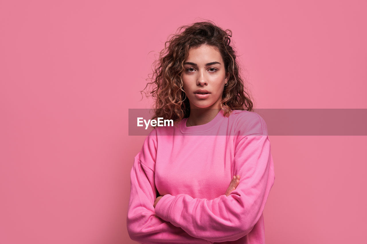 Unemotional female with curly hair standing with arms crossed looking at camera in studio with pink background