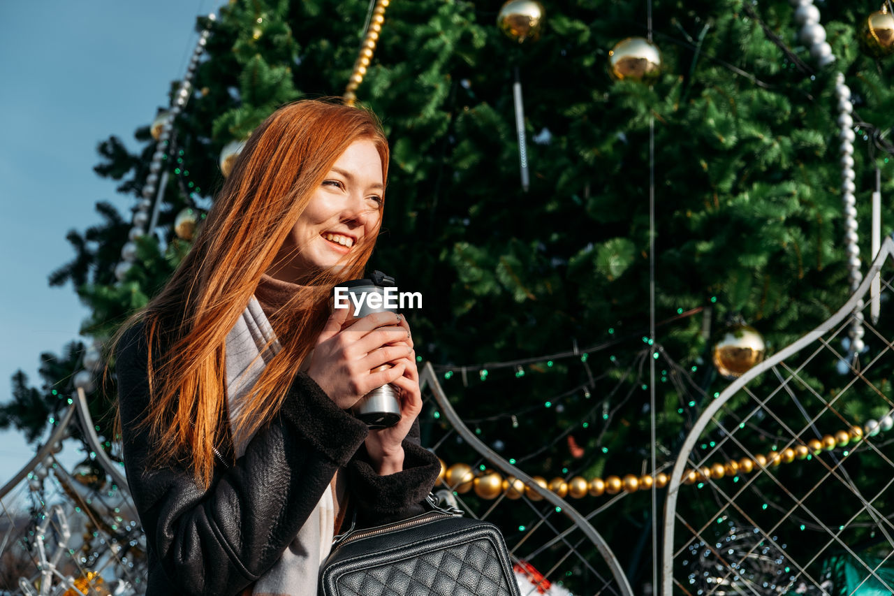 Smiling young woman looking away while holding coffee cup against trees