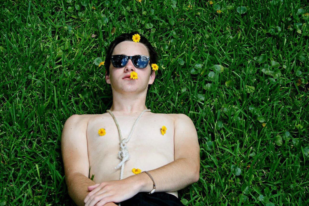 High angle view of shirtless young man wearing sunglasses with flowers lying on grassy field at park