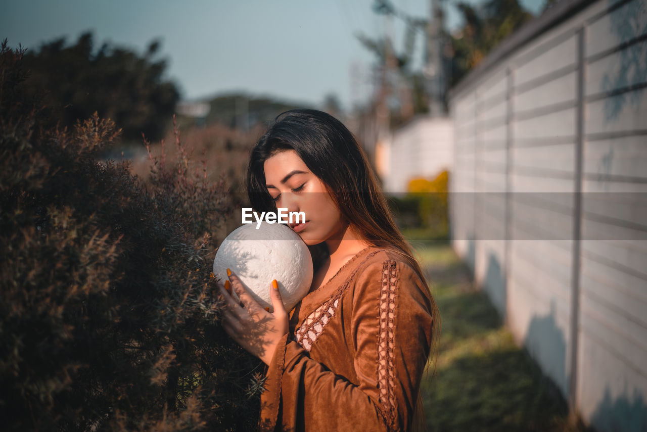 YOUNG WOMAN LOOKING AWAY WHILE STANDING AGAINST TREE