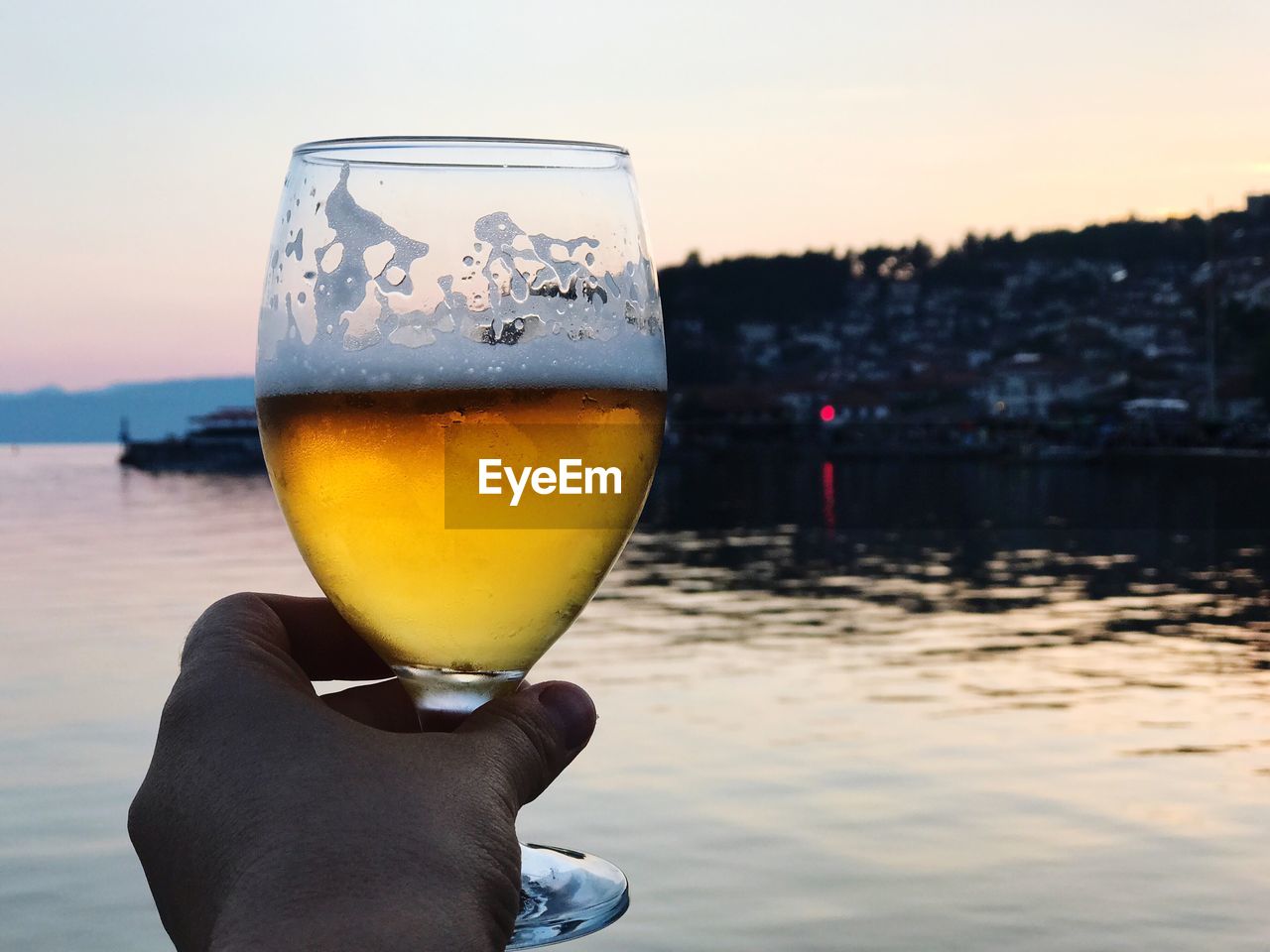 Close-up of hand holding beer glass by river at sunset