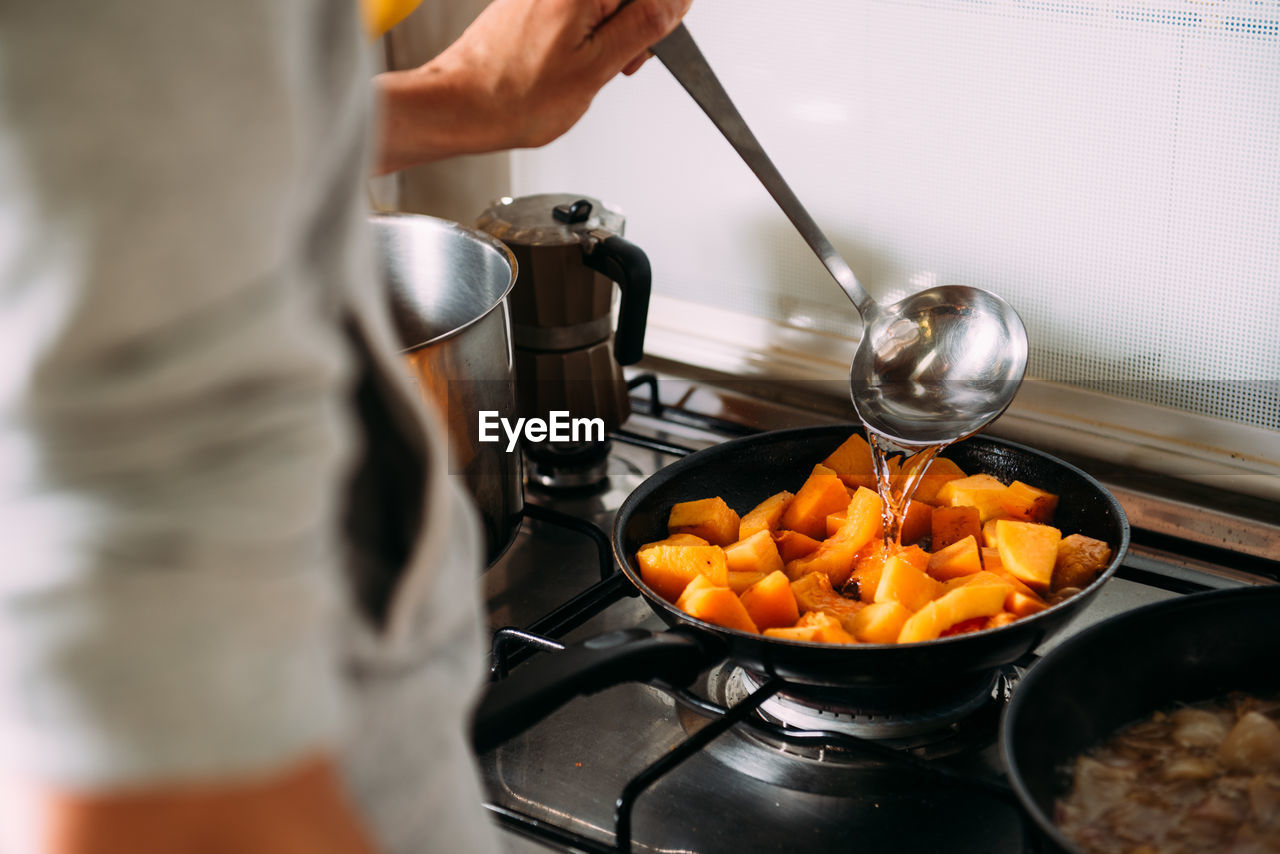 Crop anonymous woman with kitchen spoon pouring water into frying pan with chopped pumpkin while preparing cream soup