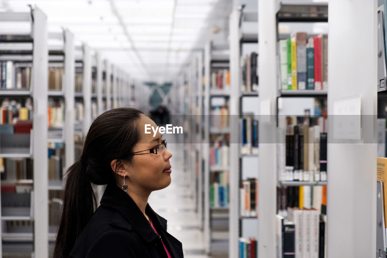 Side view of young woman looking books in shelf at library