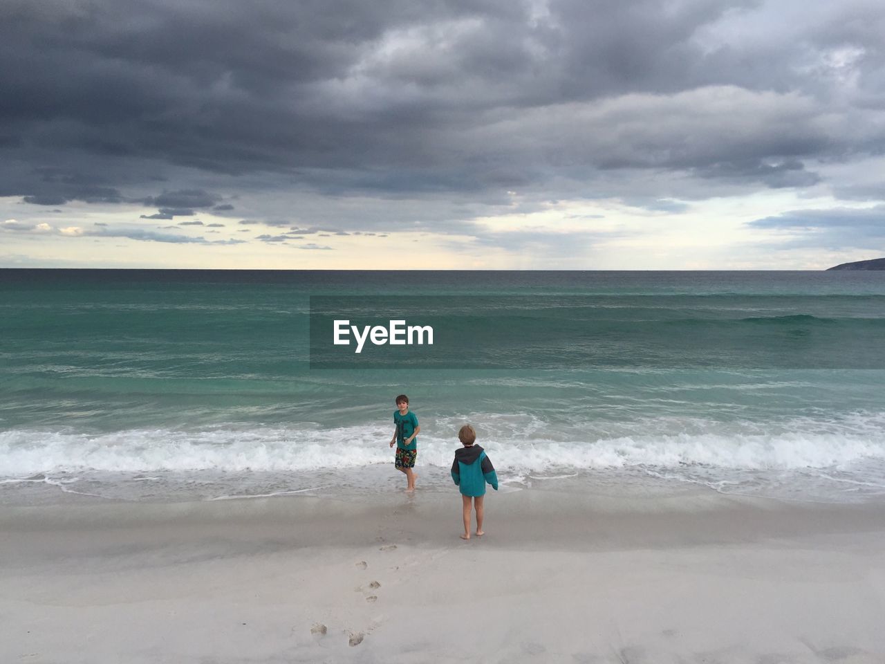 REAR VIEW OF PEOPLE AT BEACH AGAINST CLOUDY SKY