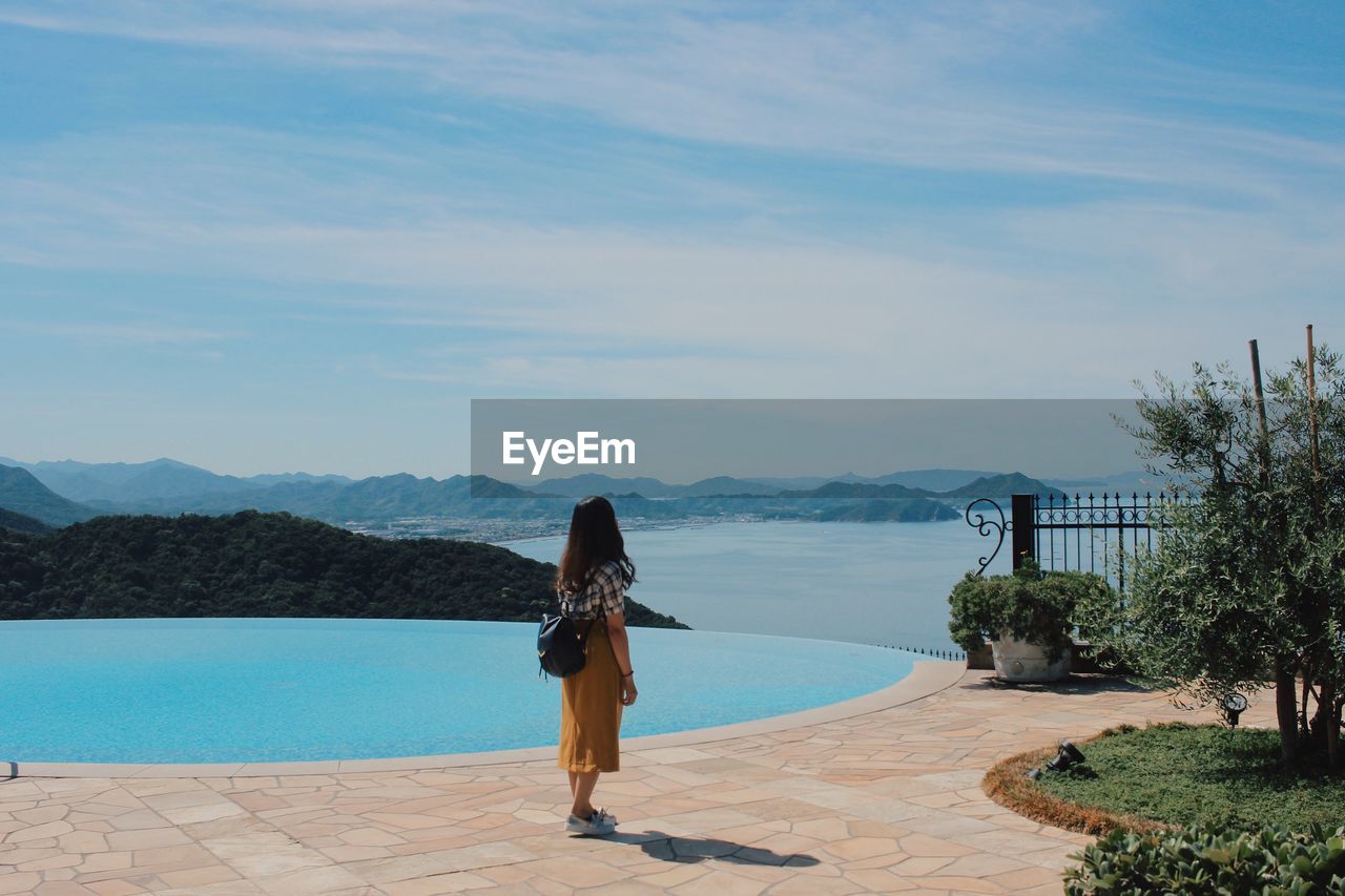 Rear view of woman standing by swimming pool against sky