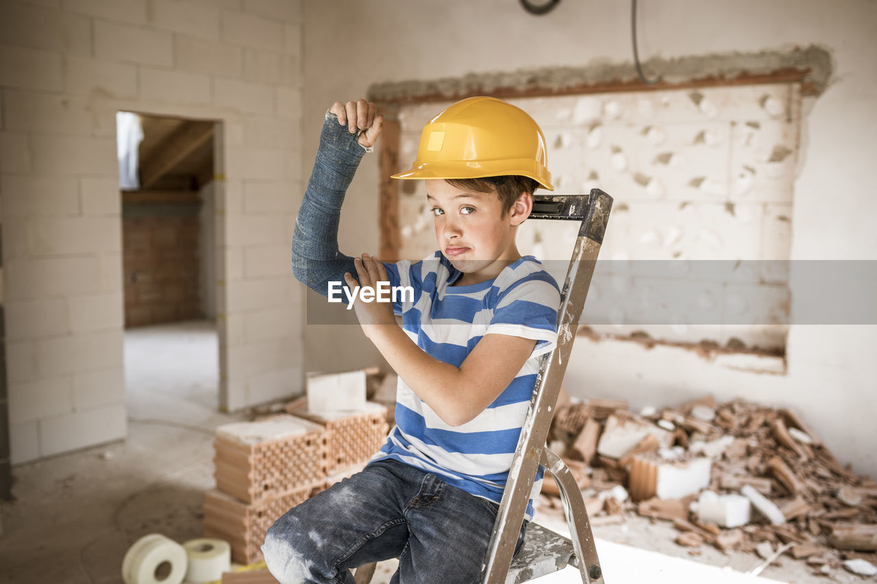 Boy flexing broken arm muscles while sitting on ladder during house renovation