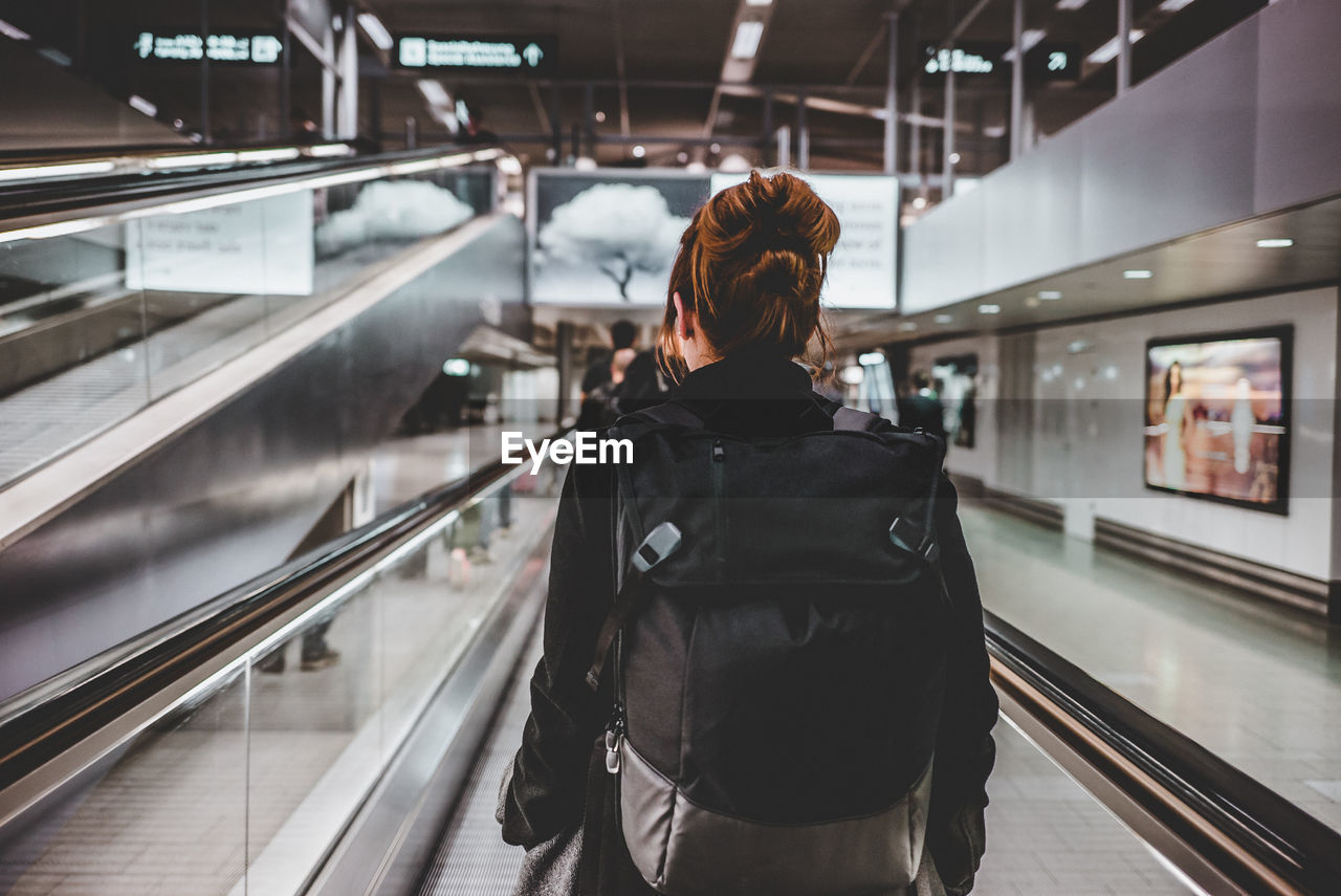 Rear view of woman with backpack standing on moving walkway at railroad station