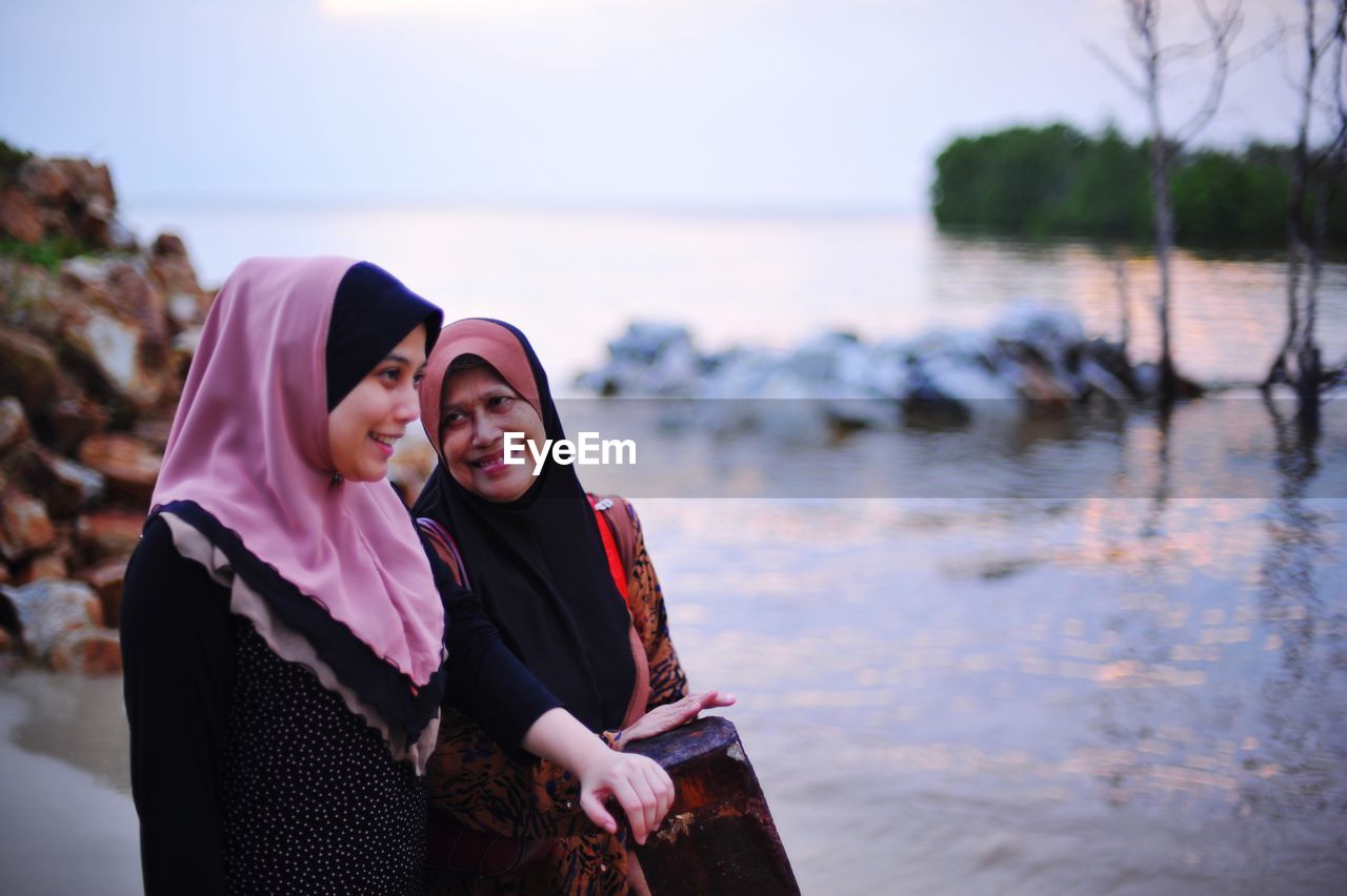 Smiling mother with daughter in hijab standing at beach against sky during sunset