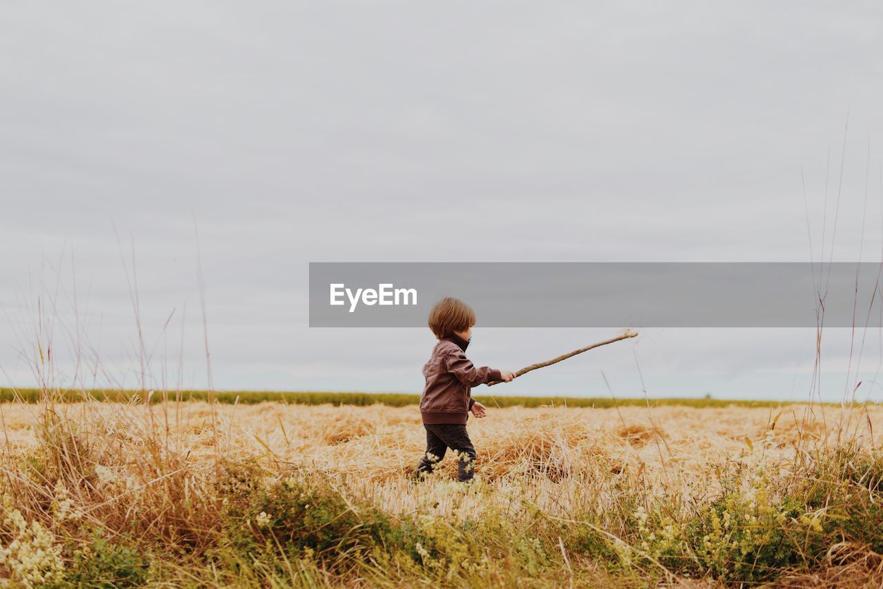 Boy holding stick while walking on grassy field against cloudy sky