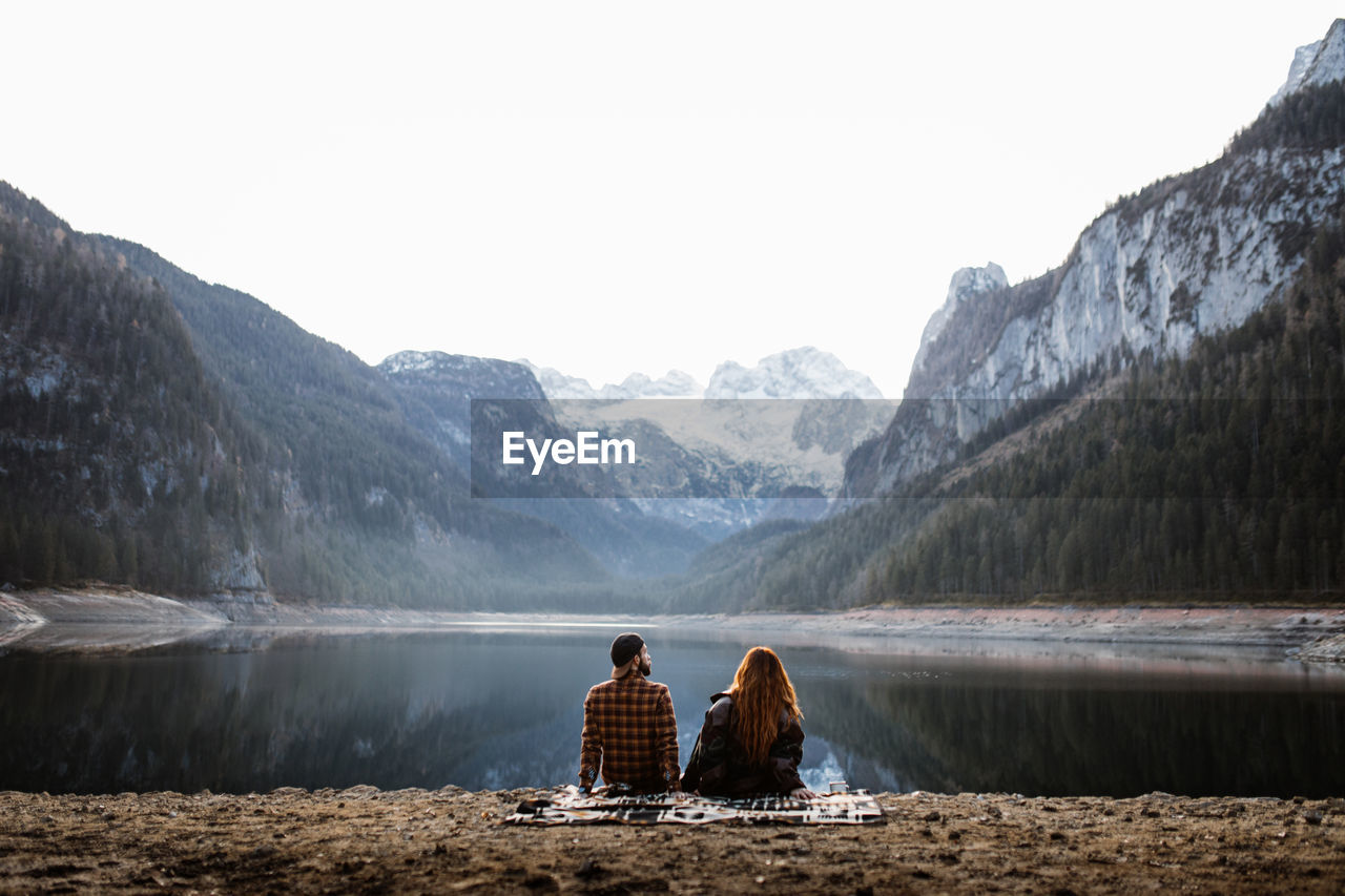 Back view of unrecognizable couple in warm clothes sitting on shore of peaceful lake and enjoying spectacular landscape of snowy mountains in autumn morning