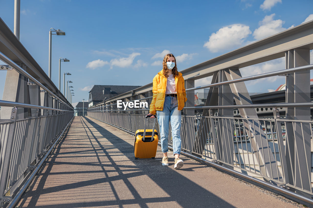 Full length of girl wearing mask walking with suitcase against sky