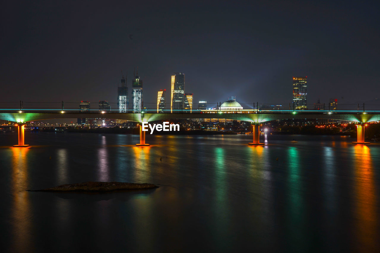 ILLUMINATED BRIDGE OVER RIVER BY BUILDINGS AT NIGHT