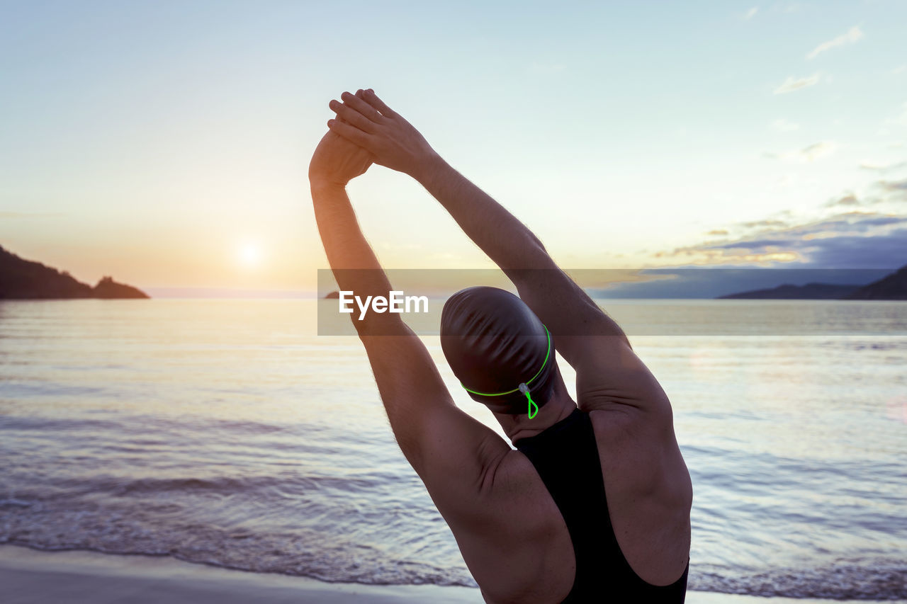 Back view of fit male swimmer in swimsuit and cap standing on seashore and stretching arms before training