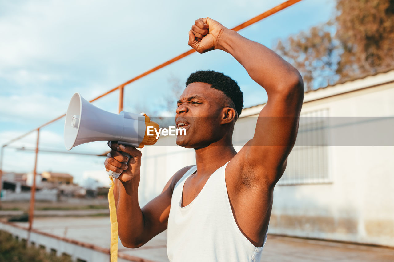 Young angry african american male in undershirt with speaker yelling with raised arm while looking at camera