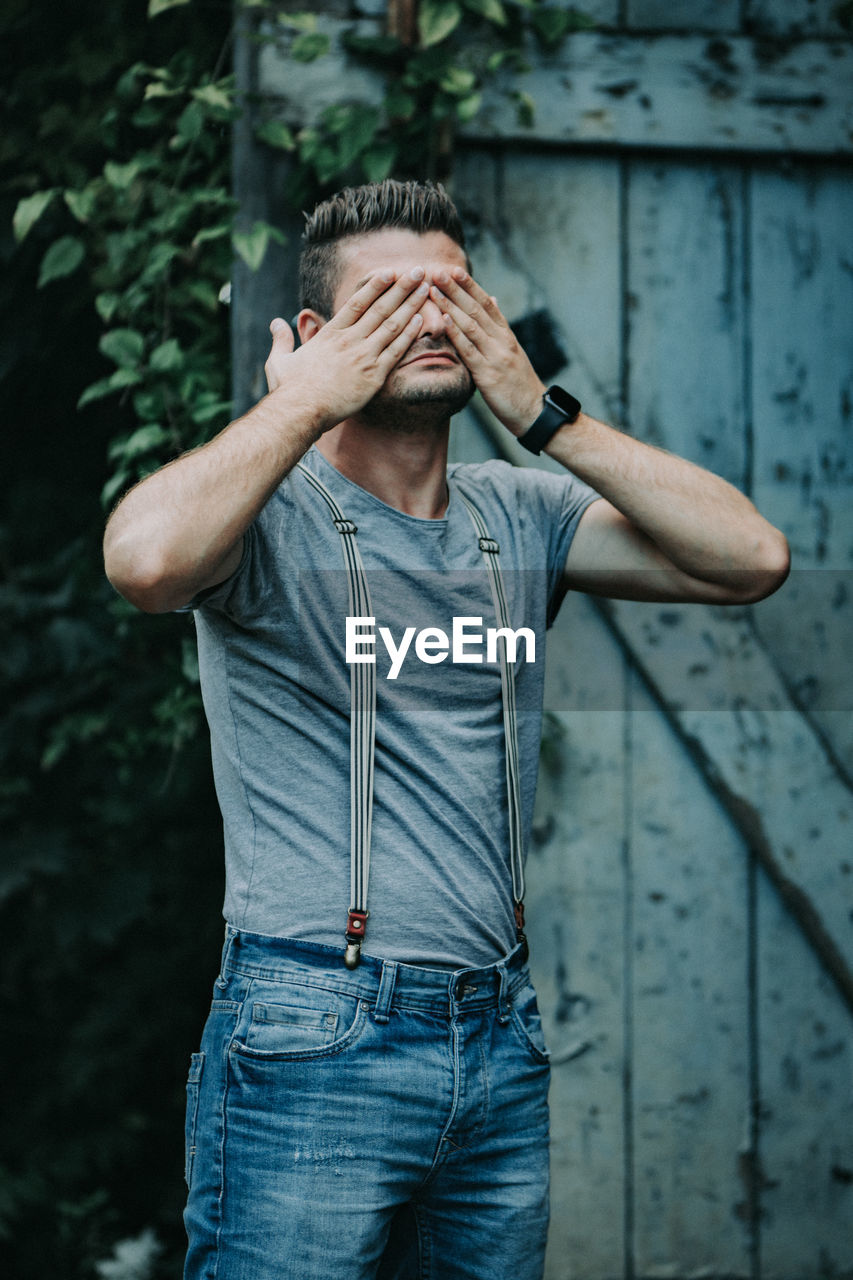 Young man with hands covering eyes standing against door