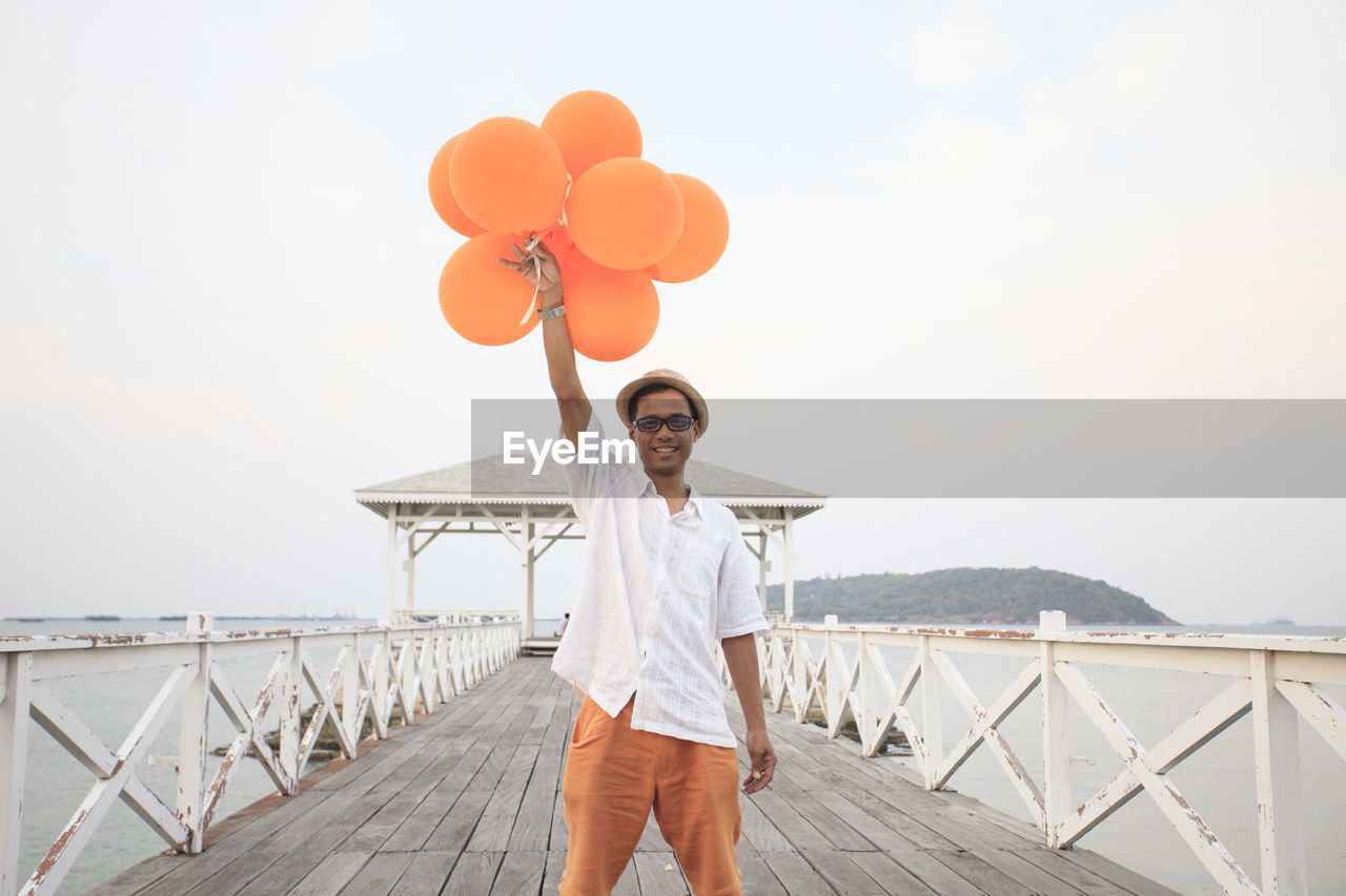 Portrait of man holding balloons while standing on pier against sky