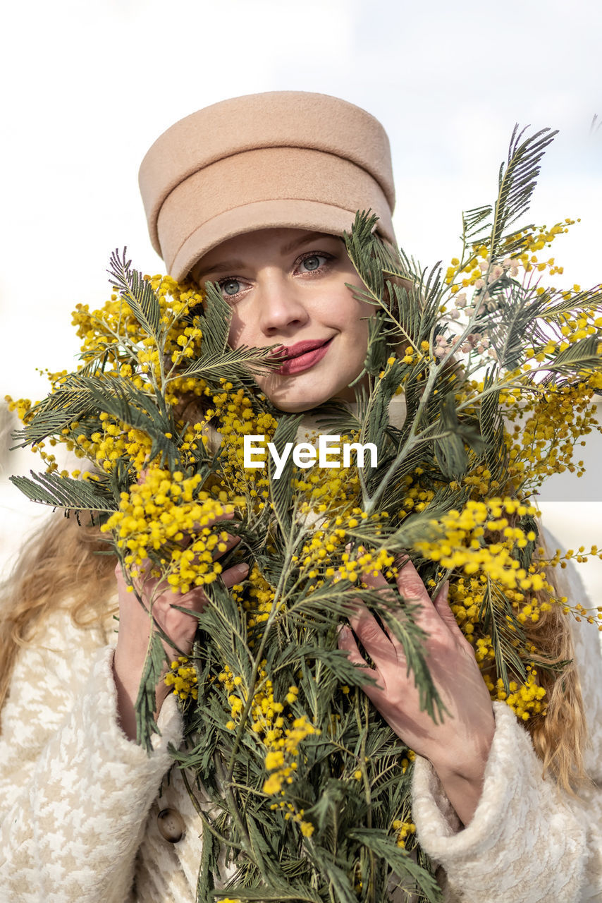 yellow, plant, one person, portrait, nature, adult, flower, clothing, flowering plant, smiling, looking at camera, hat, women, young adult, spring, happiness, emotion, holding, front view, waist up, outdoors, fashion accessory, freshness, floristry, winter, leisure activity, day, lifestyles, beauty in nature, growth, standing, cheerful, female, agriculture, floral design, sky, rural scene, tree, warm clothing