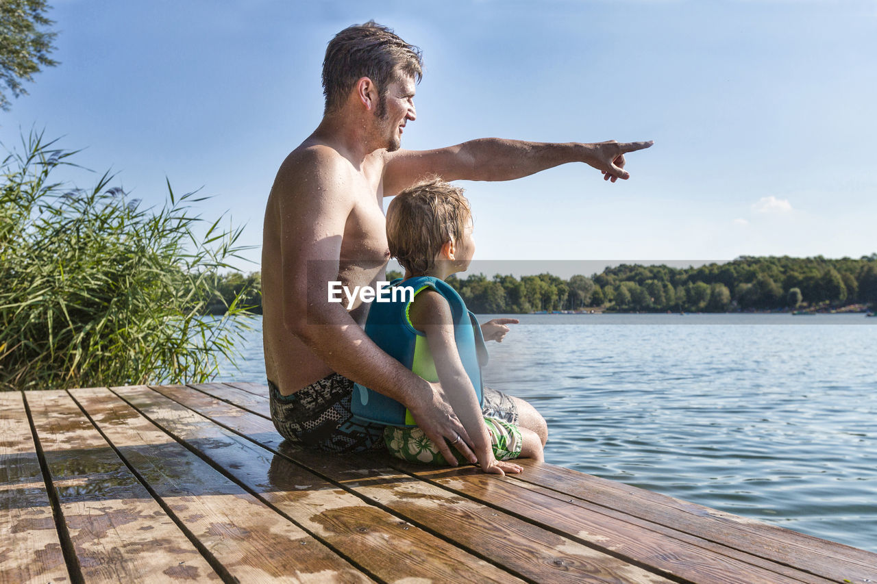 Father pointing while sitting with son on wet pier over lake against sky