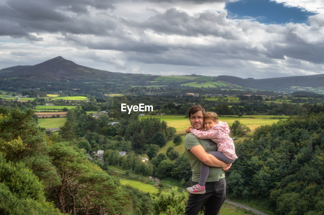 Father holding his daughter in arms with a scenic view on a valley, forest and mountain range