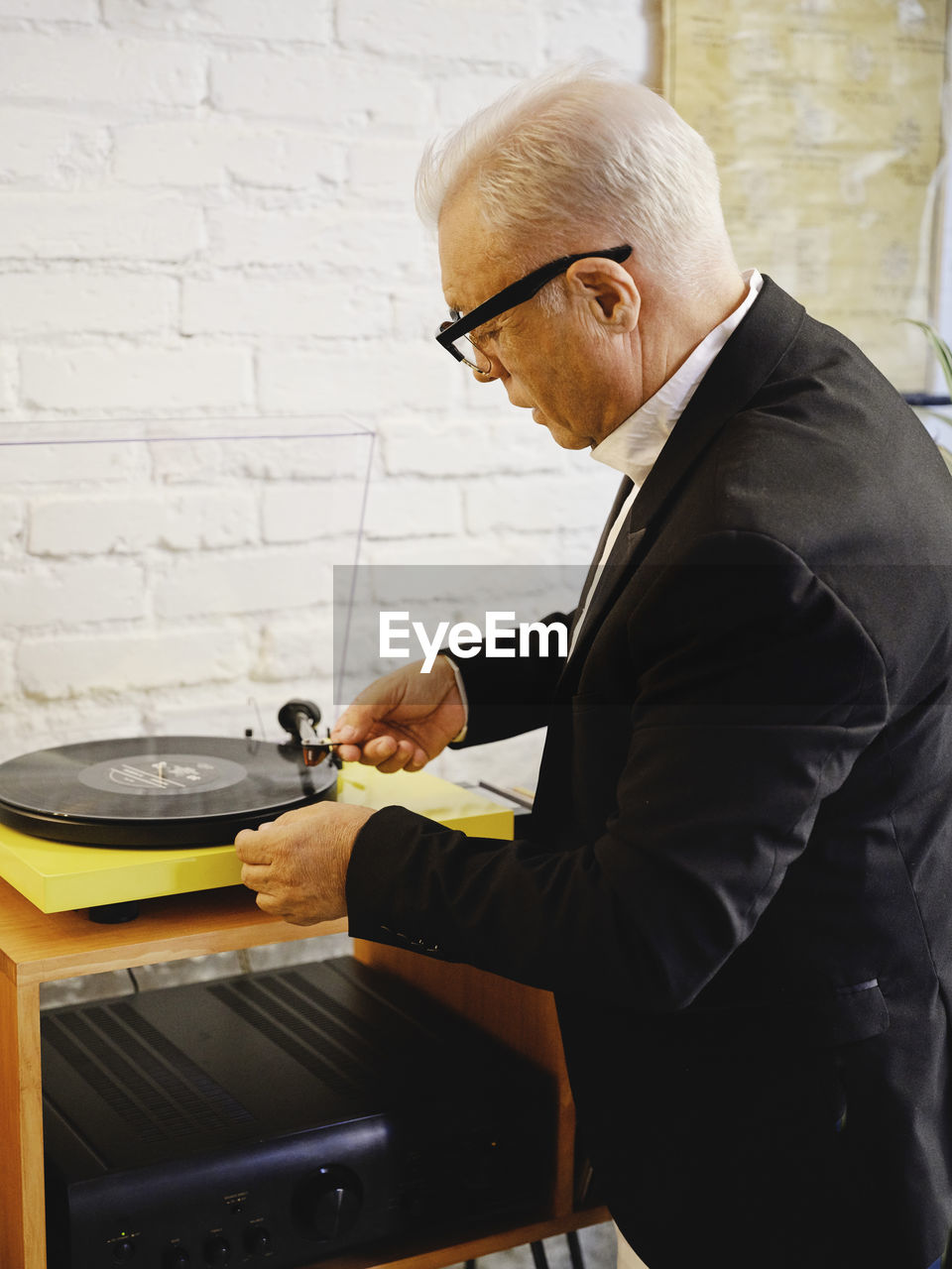Side view of senior male in elegant jacket standing near table with retro record player and putting vinyl disc for listening to music