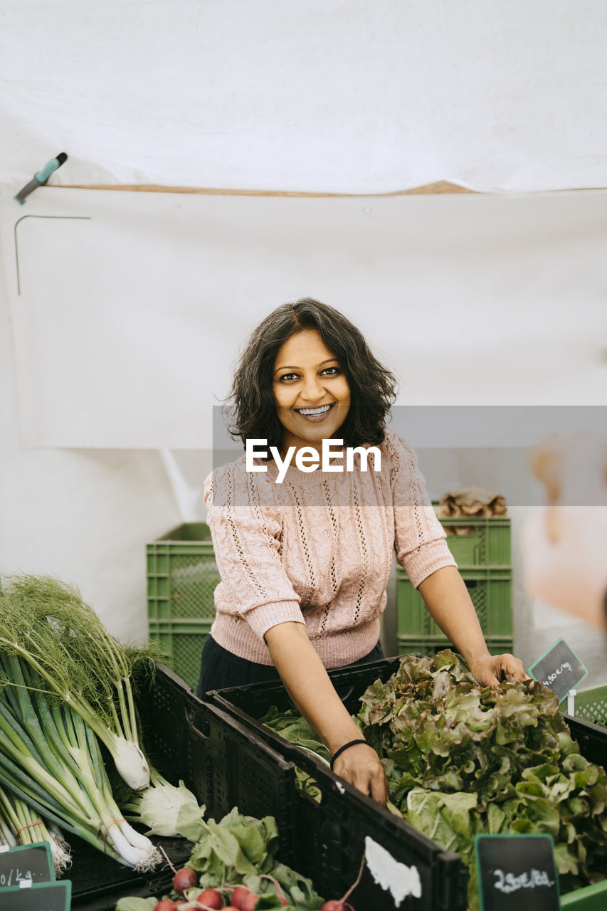 Portrait of smiling female owner standing near vegetable crate in stall at market