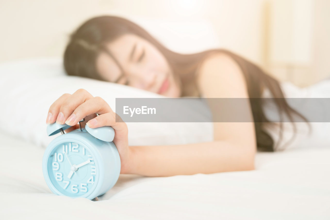 Young woman holding alarm clock while sleeping on bed