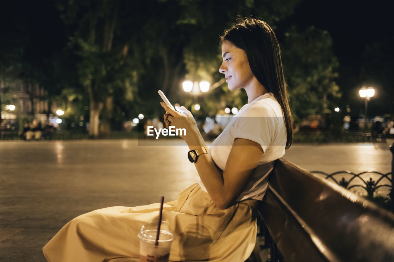 Woman using smart phone sitting on park bench at night
