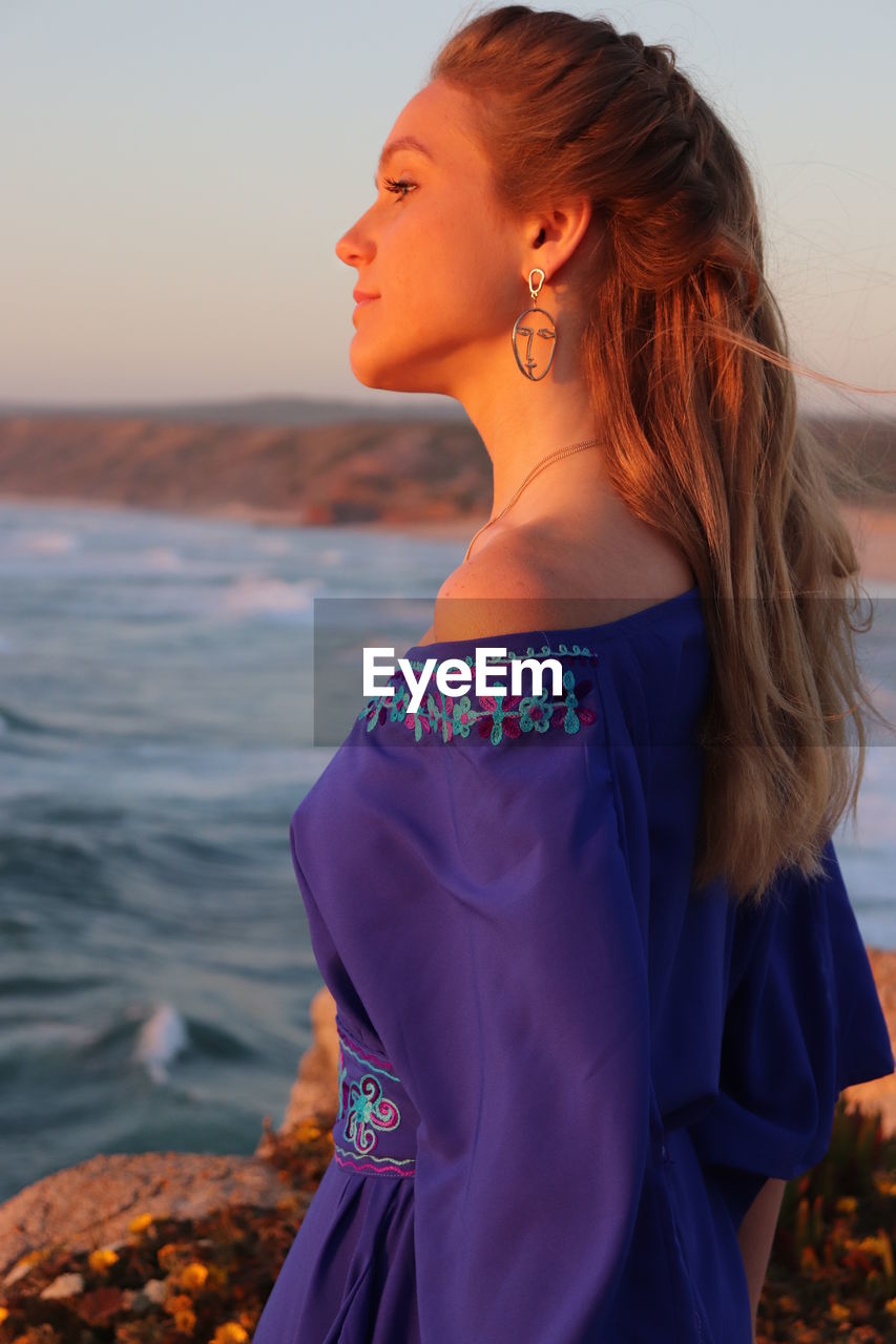 Beautiful woman standing at beach against sky during sunset